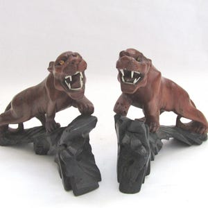 May include: Two carved wooden tiger figurines. The tigers are brown with open mouths and white teeth, perched on black, textured bases. The figurines are displayed against a white background.