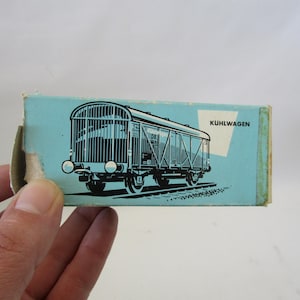 May include: A vintage blue box featuring a black and white illustration of a train car. The box has the word "KUHLWAGEN" printed on it. The box is held by a hand.