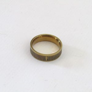 Op de afbeelding: Een goudkleurige ring met gegraveerde tekst en een kruissymbool. De ring heeft een band met gegraveerde tekst rondom de omtrek. Het kruis is gecentreerd op de band. De ring ligt op een witte ondergrond.