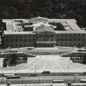 May include: A monochrome aerial view of a large building with a symmetrical design, likely a government building. The building is surrounded by a plaza and a road with vehicles, captured from above.