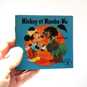 May include: A vintage children's book titled "Mickey et Wamba-Wu" featuring Mickey Mouse and another character. The cover is blue with illustrations of the characters shaking hands against an orange and yellow background. The book is held by a hand.