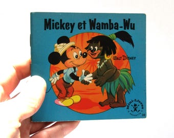 May include: A vintage children's book titled "Mickey et Wamba-Wu" featuring Mickey Mouse and another character. The cover is blue with illustrations of the characters shaking hands against an orange and yellow background. The book is held by a hand.