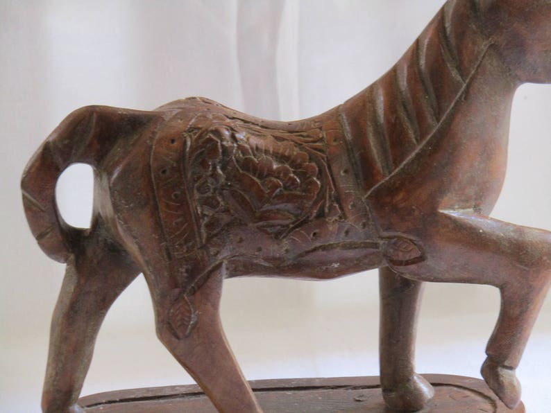 May include: A carved wooden horse figurine with intricate details. The horse is brown and has a saddle with floral designs. The figurine is standing on a wooden base.