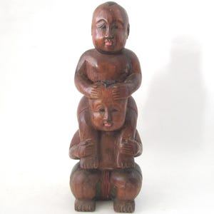 May include: A hand-carved wooden sculpture featuring two figures stacked vertically. The top figure sits on the shoulders of the bottom figure, with hands resting on the head of the lower figure. The wood has a polished, dark brown finish.