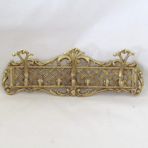 May include: An antique brass coat rack for wall mounting, featuring six hooks. The rack has an intricate openwork pattern and decorative detailing. The brass has a warm, golden colour. A vintage-style piece for hallways or bedrooms.