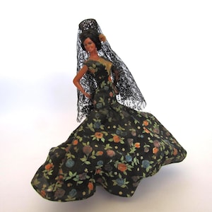 May include: A doll with dark hair wearing a black lace veil and a black floor-length dress featuring a multi-colored floral print with orange, blue, and green flowers. The doll has a gold-colored bodice detail and a small flower accessory.