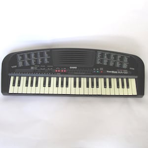 May include: A black Casio ToneBank MA-120 electronic keyboard featuring a standard layout of white and black keys, a central speaker grille, and printed control panels on both sides of the top surface with various function labels and settings.
