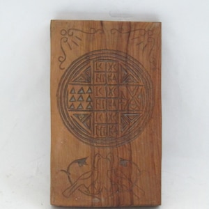 May include: A rectangular wooden carving with intricate designs. The central motif is a circular pattern with geometric shapes and symbols. The wood has a warm, natural tone, and the carvings are dark. Two bird-like figures are carved at the bottom.