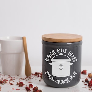 May include: Gray ceramic crock pot storage container with a wooden lid. The container has a white graphic of a crock pot with the text "Rock Out With Your Crock Out".