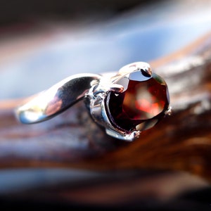 May include: A silver ring with a red gemstone. The ring is set with a round, faceted gemstone that is surrounded by a silver setting. The ring is on a wooden surface.