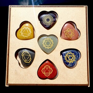 May include: Set of seven heart-shaped chakra stones in a wooden box. Each stone is a different color and has a gold chakra symbol etched on it. The stones are arranged in a circle in the box.