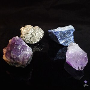 May include: Four raw crystals on a black background. A purple amethyst, a light purple amethyst, a silver pyrite, and a blue lapis lazuli.