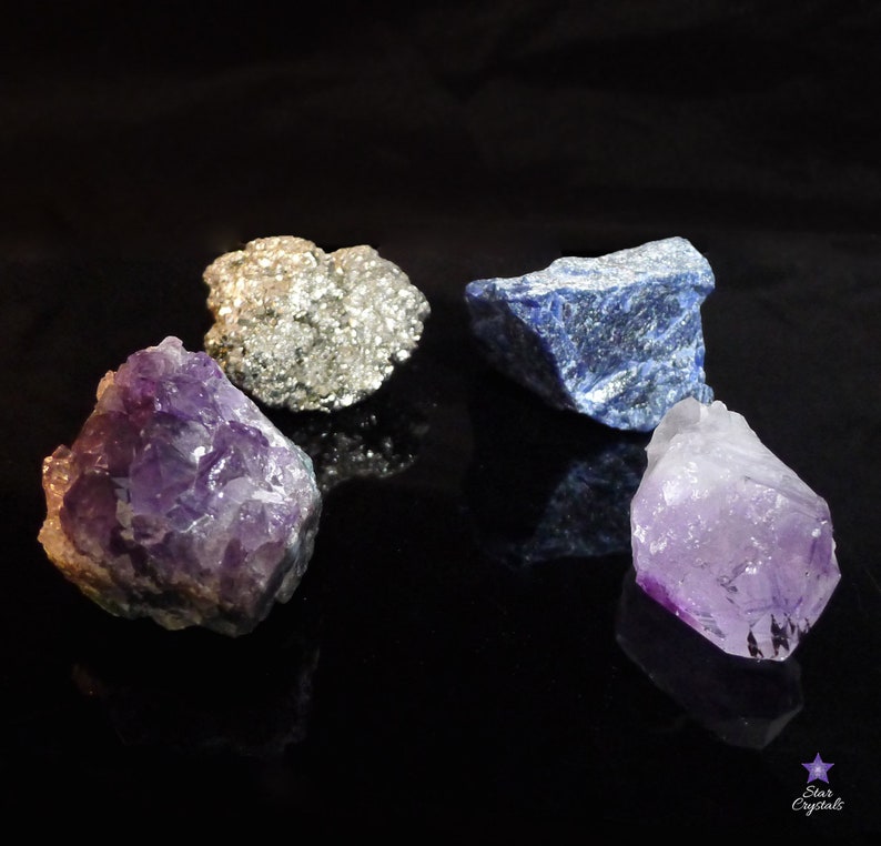 May include: Four raw crystals on a black background. A purple amethyst, a light purple amethyst, a silver pyrite, and a blue lapis lazuli.