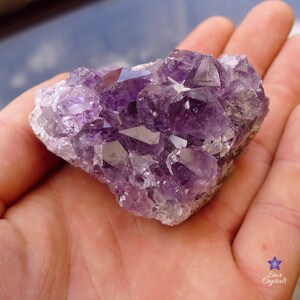 May include: A cluster of amethyst crystals in various shades of purple. The crystals are clustered together in a natural formation, with some points and some flat surfaces. The crystals are translucent and have a slight sparkle.