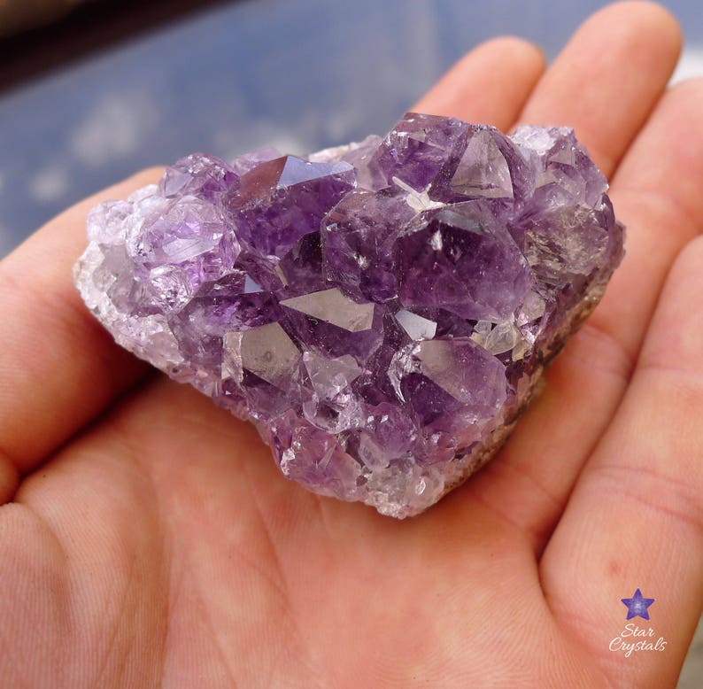 May include: A cluster of amethyst crystals in various shades of purple. The crystals are clustered together in a natural formation, with some points and some flat surfaces. The crystals are translucent and have a slight sparkle.