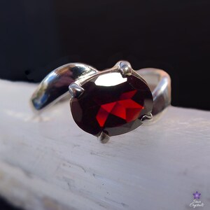 May include: A silver ring with a red garnet gemstone. The ring has a curved band and the gemstone is set in a prong setting.