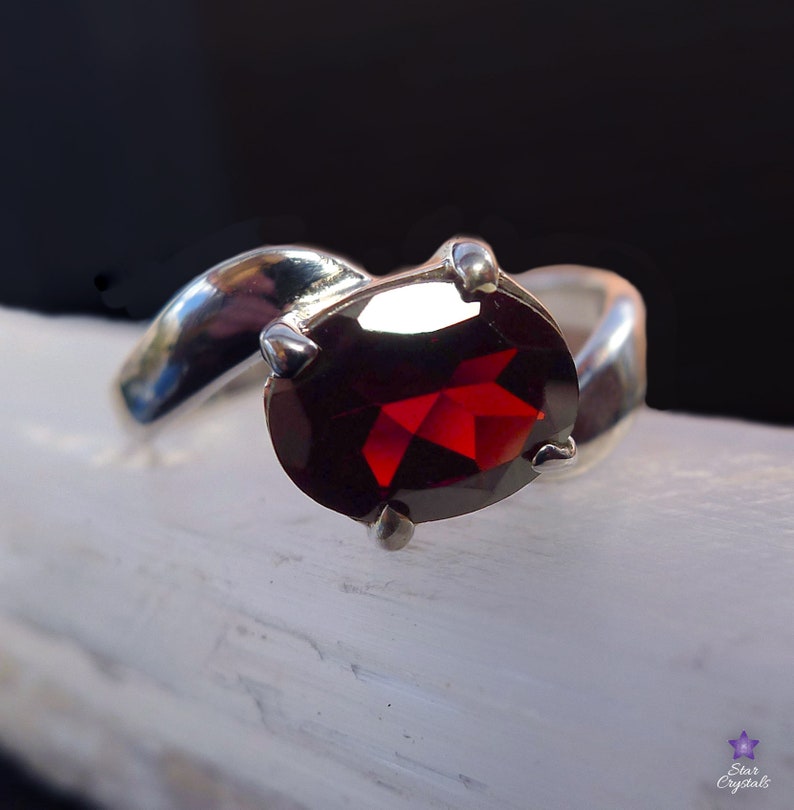 May include: A silver ring with a red garnet gemstone. The ring has a curved band and the gemstone is set in a prong setting.