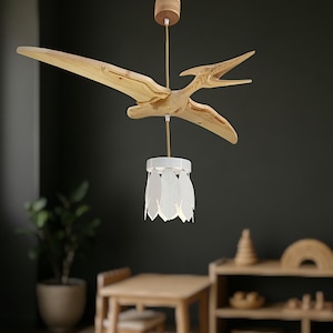 May include: A wooden Pterodactyl-shaped pendant light. The dinosaur is made of light-coloured wood, with a white lampshade featuring leaf-shaped cutouts. The fixture hangs from a wooden ceiling mount.