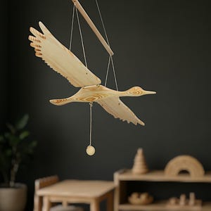 May include: A wooden bird mobile with outstretched wings, suspended by strings. The bird is made of light-colored wood with visible grain, and a small wooden ball hangs below. The background is a dark wall with a glimpse of furniture.
