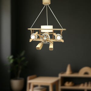 May include: A ceiling pendant light shaped like a biplane, constructed from light-toned wood. The aeroplane features three circular light fixtures with white bulbs, a central cylindrical light, and wooden propellers, all suspended by white cords.