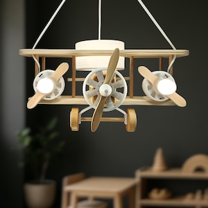 May include: A hanging light fitting shaped like a biplane, constructed from light-toned wood and white components. The design features three wooden propellers, two illuminated circular light bulbs, and a central cylindrical white lampshade.
