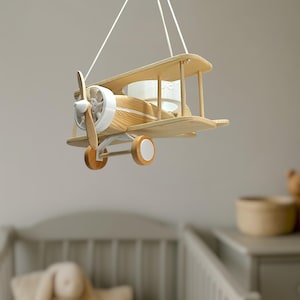 May include: A hanging biplane model crafted from light-toned wood with white accents. The aeroplane features a front propeller, two wings connected by vertical struts, and a landing gear assembly with two wheels, all suspended by white strings.