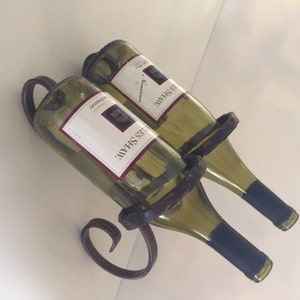 May include: A wrought iron wine bottle holder with two green wine bottles. The bottles are held in place by a dark brown metal frame. The bottles have white labels with red and black text.