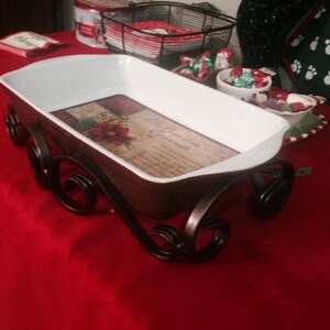 May include: A white rectangular serving dish with a floral design, set on a dark brown metal sleigh base. The dish is on a red tablecloth, suggesting a festive theme. The base has decorative scrollwork.