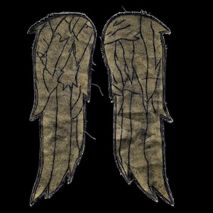 May include: A pair of embroidered patches shaped like wings, featuring a textured, muted gold fabric base with dark black thread detailing that outlines the individual feathers and the outer edges of each wing against a black background.