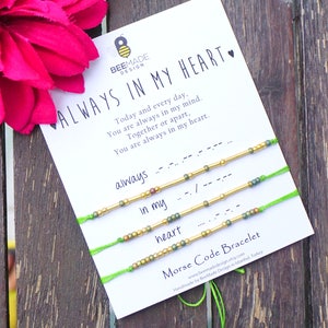 May include: A set of three green friendship bracelets with gold beads and a card that reads "Always in my heart." The card has a quote about always being in someone's mind and heart. The bracelets are made with Morse code.