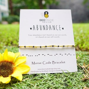 May include: A gold and black beaded bracelet with the word "ABUNDANCE" written on a white card. The card also includes the text "True abundance isn't based on our net worth, it's based on our self worth." and "Morse Code Bracelet".
