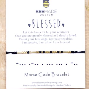 May include: A black string bracelet with gold and black beads spelling out "blessed" in Morse code. The bracelet is on a white card with the text "BeeMade Design" and "Blessed" in black text.