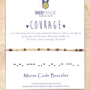 Könnte beinhalten: Ein gold- und braunes Perlenarmband mit einer Karte, auf der "COURAGE" in lila Schrift steht. Die Karte enthält auch den Morsecode für das Wort "COURAGE" und den Text "Lass dieses Armband dich daran erinnern, dass es Mut braucht, um aufzuwachsen und zu dem Menschen zu werden, der du wirklich bist. Sei mutig, hab Mut, sei freundlich." Die Karte enthält auch die Website-Adresse "www.beemadedesign.etsy.com" und den Text "Handgefertigt von BeeMade Design in Istanbul, Türkei."