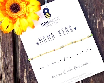 Könnte beinhalten: Eine weiße Karte mit schwarzem Text, der "BEEMADE DESIGN" und "MAMA BEAR" mit einem kleinen Herzsymbol lautet. Ein gelbes und grünes Perlenarmband liegt auf der Karte, darunter der Morsecode für "mama". Die Karte liegt auf einer Holzoberfläche mit einer gelben Blume.