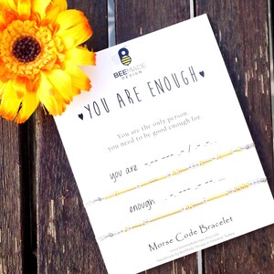 Op de afbeelding: Een witte kaart met de tekst "YOU ARE ENOUGH" en een bijenlogo. De kaart bevat ook de tekst "You are the only person you need to be good enough for" en "you are" en "enough" geschreven in morsecode. De kaart ligt op een houten oppervlak met een gele bloem.