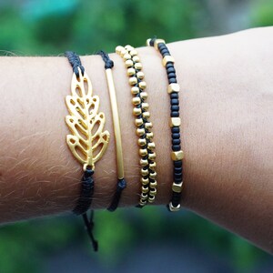 May include: A set of four bracelets featuring a gold leaf charm, a gold bar, gold beads, and black square beads. The bracelets are on a person's wrist.
