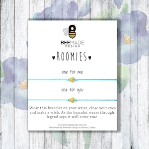 May include: Two blue friendship bracelets with gold bee charms. The bracelets are displayed on a white card with the text "ROOMIES" and "one for me" and "one for you".