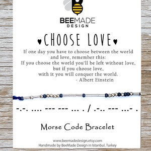 Pode incluir: Um bracelete azul e prateado com um cordão preto. O bracelete é um bracelete de código Morse com a mensagem "... /.-" que se traduz como "Escolha o amor". O cartão também inclui uma citação de Albert Einstein: "Se um dia você tiver que escolher entre o mundo e o amor, lembre-se disso: Se você escolher o mundo, você ficará sem amor, mas se você escolher o amor, com ele você conquistará o mundo."