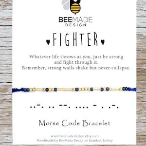 Pode incluir: Um bracelete de contas azul e dourado com um cartão que diz "FIGHTER" e "Bracelete de código Morse". O cartão também inclui o texto "O que a vida te lançar, apenas seja forte e lute por isso. Lembre-se, paredes fortes tremem, mas nunca caem."