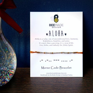 Könnte beinhalten: Eine weiße Karte mit den Worten "BeeMade Design" und "Aloha" in schwarzer Schrift. Die Karte enthält auch eine Definition des Wortes Aloha und ein kleines orangefarbenes und silbernes Perlenarmband mit dem Wort Aloha in Morsecode. Die Karte lautet "Morsecode-Armband" und enthält die Website-Adresse "www.beemadedesign.etsy.com".