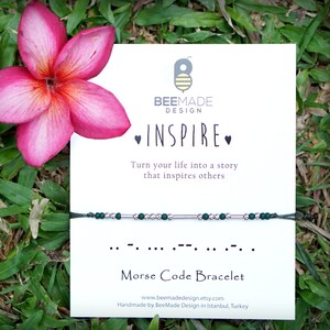 May include: A green and silver bracelet with the word "INSPIRE" written in Morse code. The bracelet is on a white card with the BeeMade Design logo and the text "Turn your life into a story that inspires others".