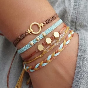 May include: A collection of handmade bracelets in various styles and colors. The bracelets include woven, beaded, and macrame designs, featuring gold-colored accents and a mix of brown, blue, and tan hues. The bracelets are displayed on a wrist.