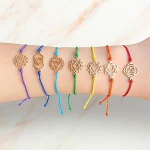 Può includere: Una collezione di sette braccialetti chakra colorati. Ogni braccialetto presenta un ciondolo color oro con un design unico, infilato su un cordino nel colore corrispondente dello spettro dei chakra: viola, blu, turchese, verde, giallo, arancione e rosso.