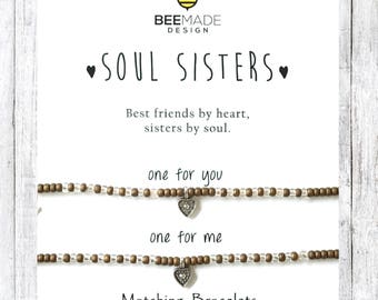 Puede incluir: Dos pulseras a juego con dijes de corazón, una para ti y una para mí. Las pulseras están hechas de cuentas marrones y blancas y tienen un dije de corazón plateado. El texto en la tarjeta dice "Soul Sisters" y "Mejores amigas de corazón, hermanas de alma."