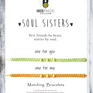 Pu&ograve; includere: Due braccialetti dell'amicizia con il testo "Soul Sisters" e "Best friends by heart, sisters by soul." I braccialetti sono verde e arancio con perline dorate. Il testo "one for you" e "one for me" &egrave; sopra i braccialetti.