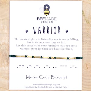 Puede incluir: Un brazalete de cuentas verdes y doradas con la palabra "WARRIOR" escrita en código Morse. La tarjeta dice: "La mayor gloria de la vida no reside en no caer nunca, sino en levantarse cada vez. Que este brazalete te recuerde que eres un guerrero, más fuerte de lo que jamás has sido." También dice "Pulsera de código Morse" e incluye la dirección del sitio web www.beemadedesign.etsy.com. La tarjeta está hecha a mano por BeeMade Design en Estambul, Turquía.