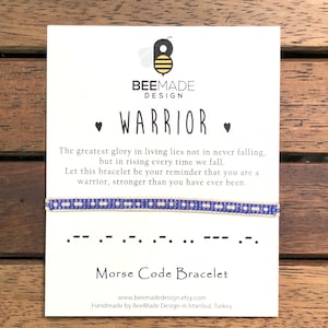 May include: A Morse code bracelet with a blue and white beaded design, displayed on a white card. The card features the word "WARRIOR" and a motivational quote. The BeeMade Design logo is visible.
