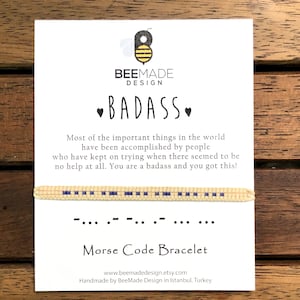 May include: A white card with the text "BEEMADE DESIGN BADASS" and a bee logo. The card also includes the text "Most of the important things in the world have been accomplished by people who have kept on trying when there seemed to be no help at all. You are a badass and you got this!" and a yellow and blue beaded bracelet with the morse code for "BADASS" on it. The card also includes the text "Morse Code Bracelet" and the website address "www.beemadedesign.etsy.com". The card also includes the text "Handmade by BeeMade Design in Istanbul, Turkey".