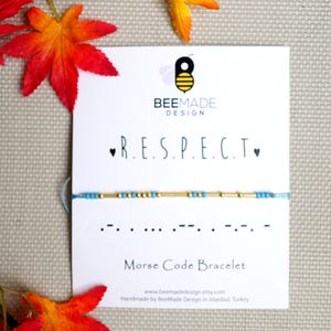 Peut inclure: Un bracelet en perles bleues et dorées avec le mot "RESPECT" écrit en code Morse. Le bracelet est sur une carte blanche avec le logo BeeMade Design et le texte "Bracelet en code Morse".
