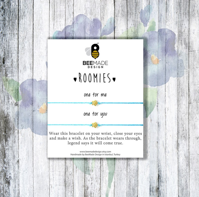May include: Two blue friendship bracelets with gold bee charms. The bracelets are displayed on a white card with the text "ROOMIES" and "one for me" and "one for you".